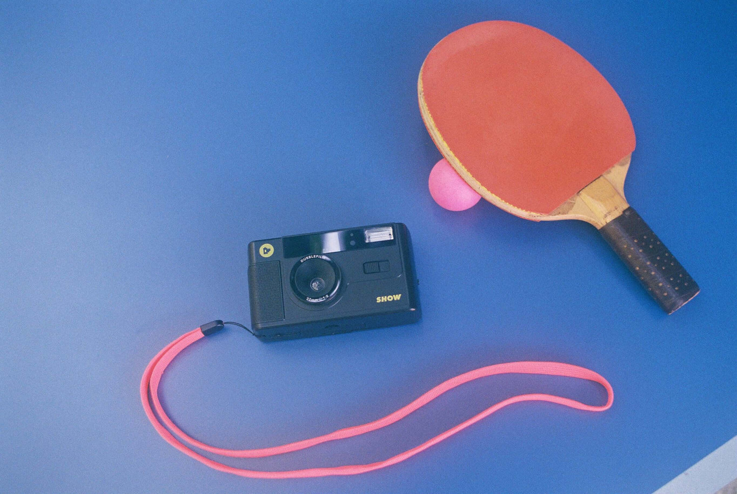 Dubblefilm SHOW 35mm Reusable Camera (Black) on blue surface with pink strap and table tennis paddle