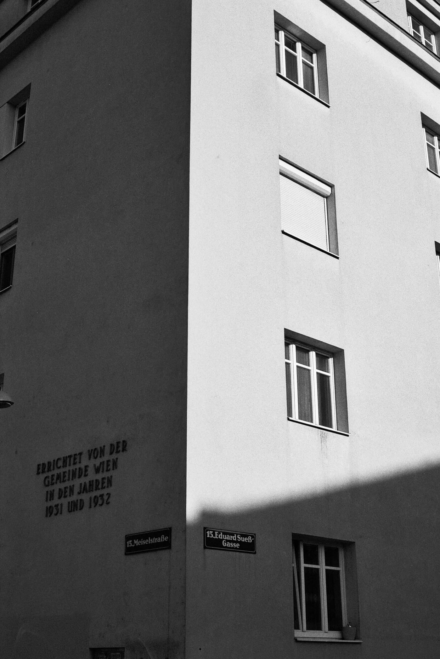 Black and white photograph of a building corner with street signs and shadows.
