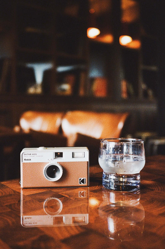 Kodak Ektar H35 Half Frame Film Camera (Brown) next to a glass of water on a wooden table.