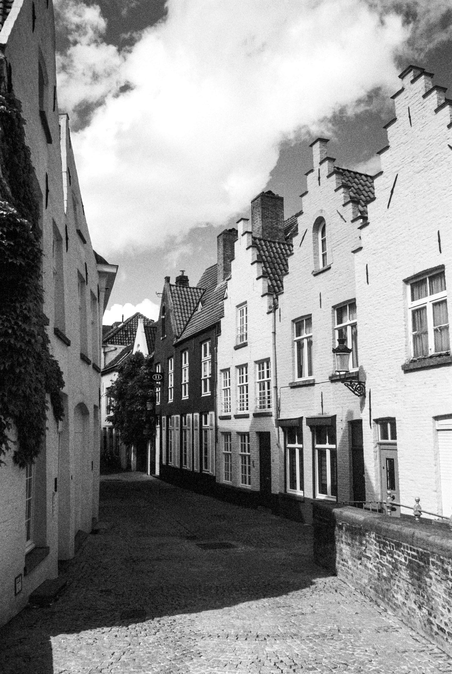 Black and white image of a narrow street with historic houses and a cloudy sky, shot on Ilford SFX 200.