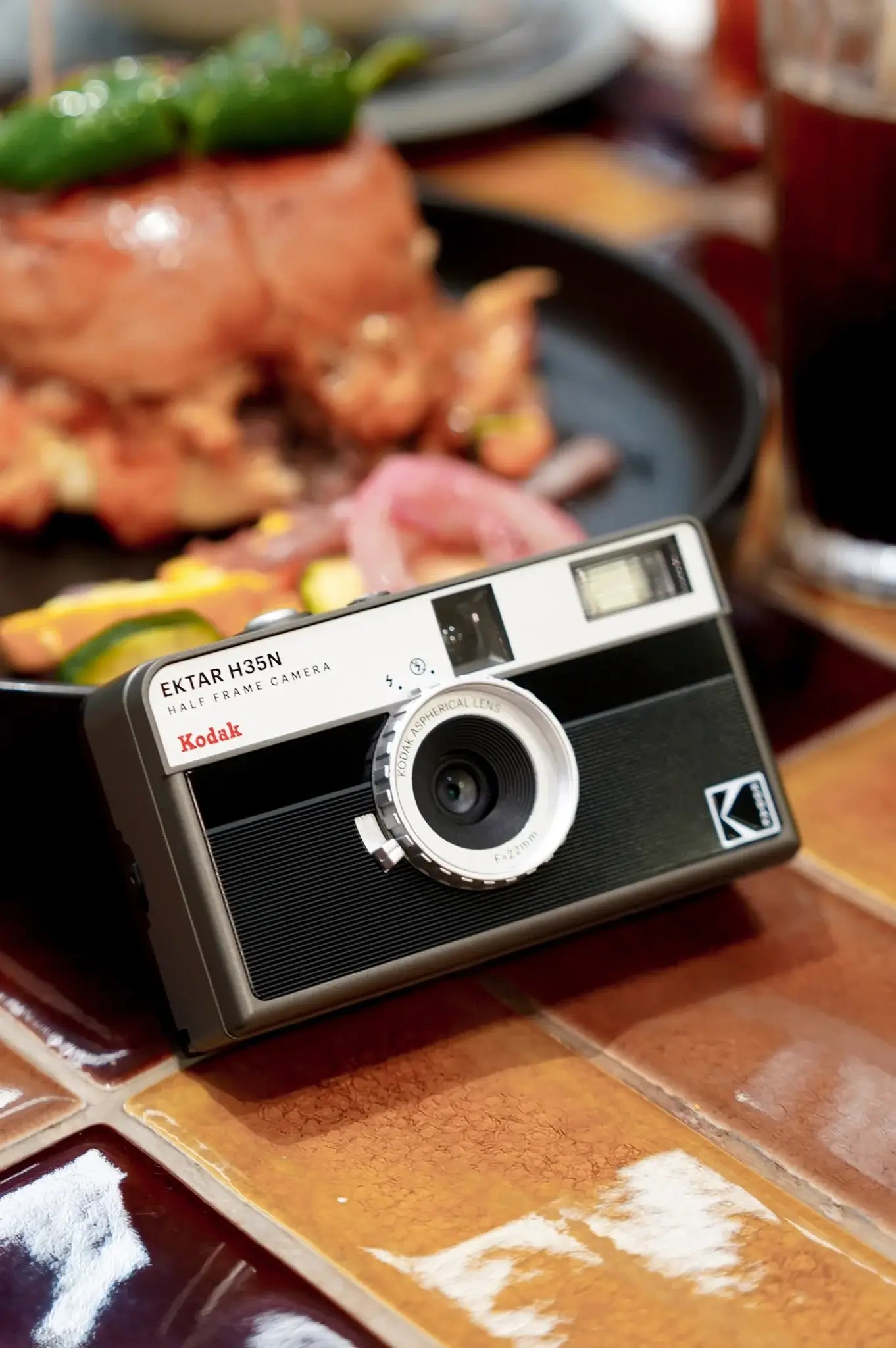 Kodak Ektar H35N Half Frame Film Camera (Striped Black) placed on a table with food in the background.
