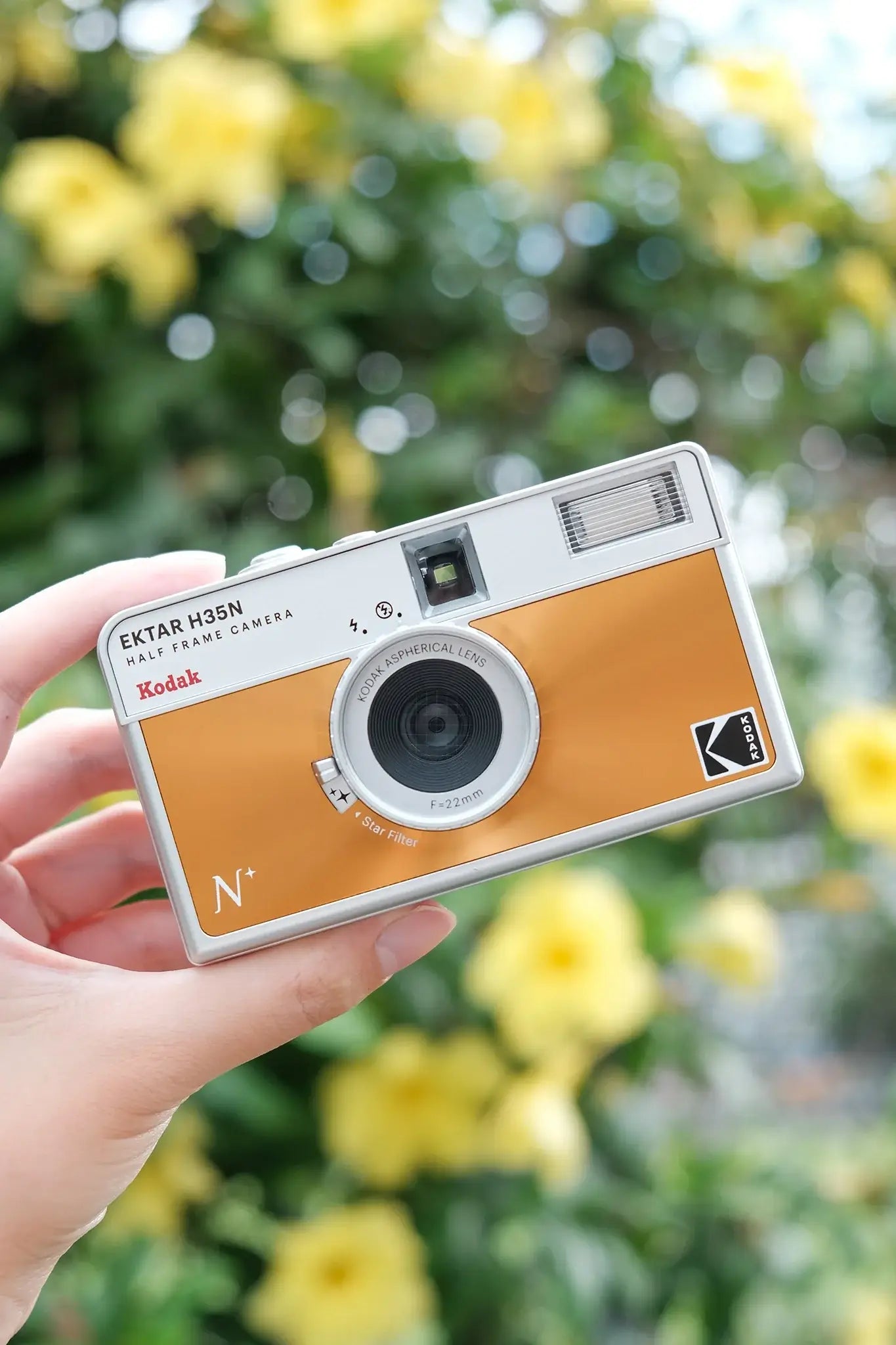 Kodak Ektar H35N Half Frame Film Camera (Orange) held in hand against a floral backdrop.