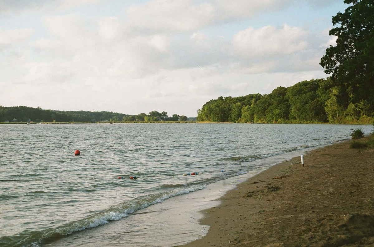 A peaceful lakeside view with a sandy beach, trees lining the shore, and buoy markers in calm water, shot on Kodak Eastman UltraMax 400.