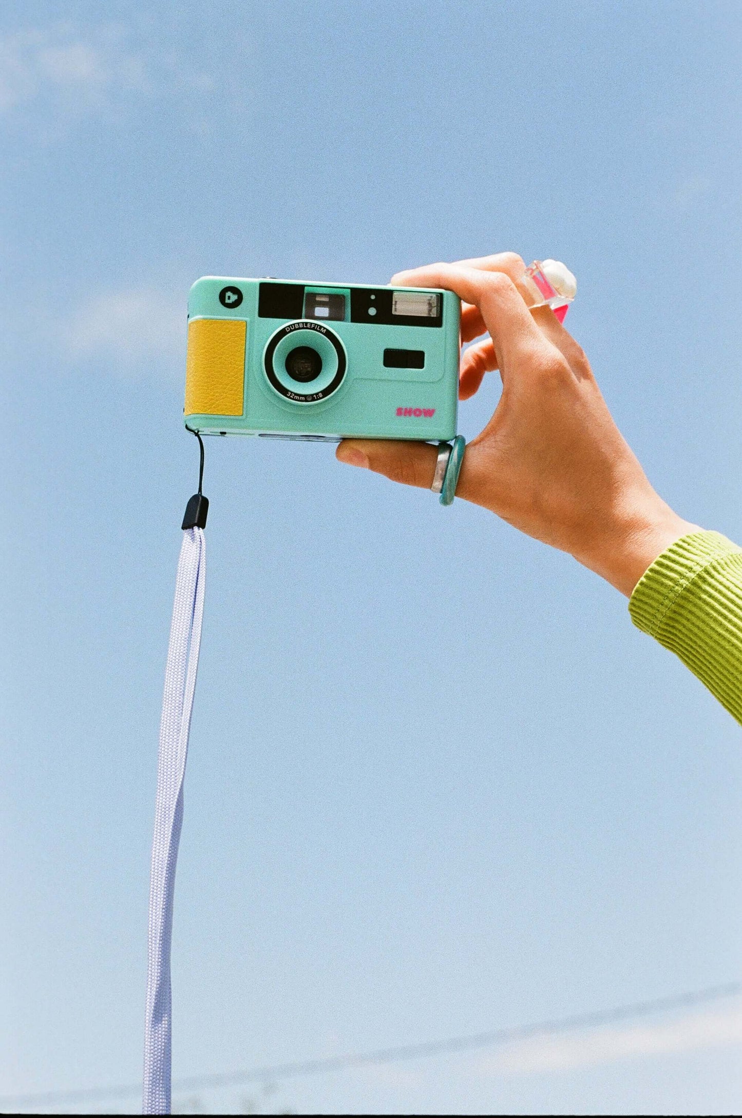Dubblefilm SHOW 35mm Reusable Camera (Turquoise) with built-in flash, turquoise body, hand holding against blue sky, includes strap and case