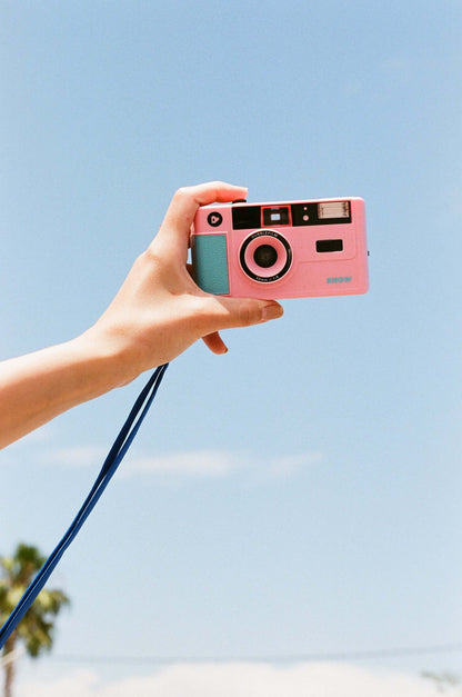 Dubblefilm SHOW 35mm Reusable Camera (Pink) held up for photo capture with sky background, built-in flash, pink housing, and strap — perfect for on-the-go photography.