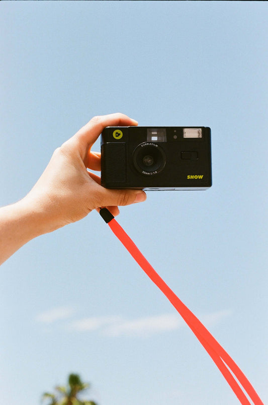 Dubblefilm SHOW 35mm Reusable Camera (Black) held in hand with red strap against blue sky