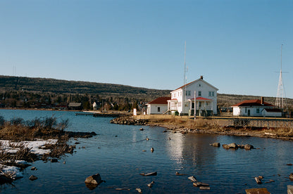 Scenic view of waterfront house with red roofs near calm water and rocky shoreline under clear blue sky, shot on Fujifilm 200.