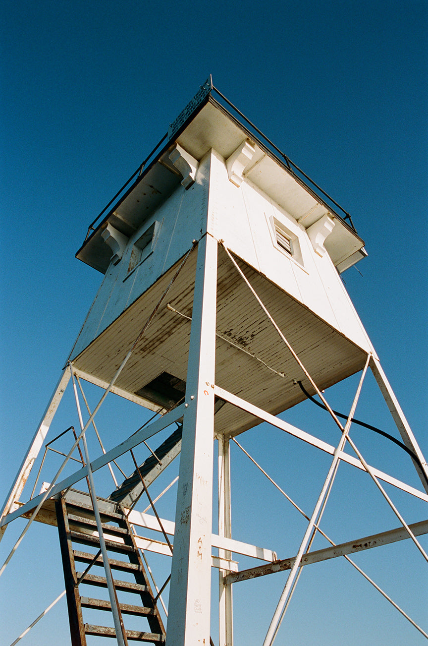 White lookout tower with stairs against a clear blue sky, showcasing its structure and height, shot on Fujifilm 200.