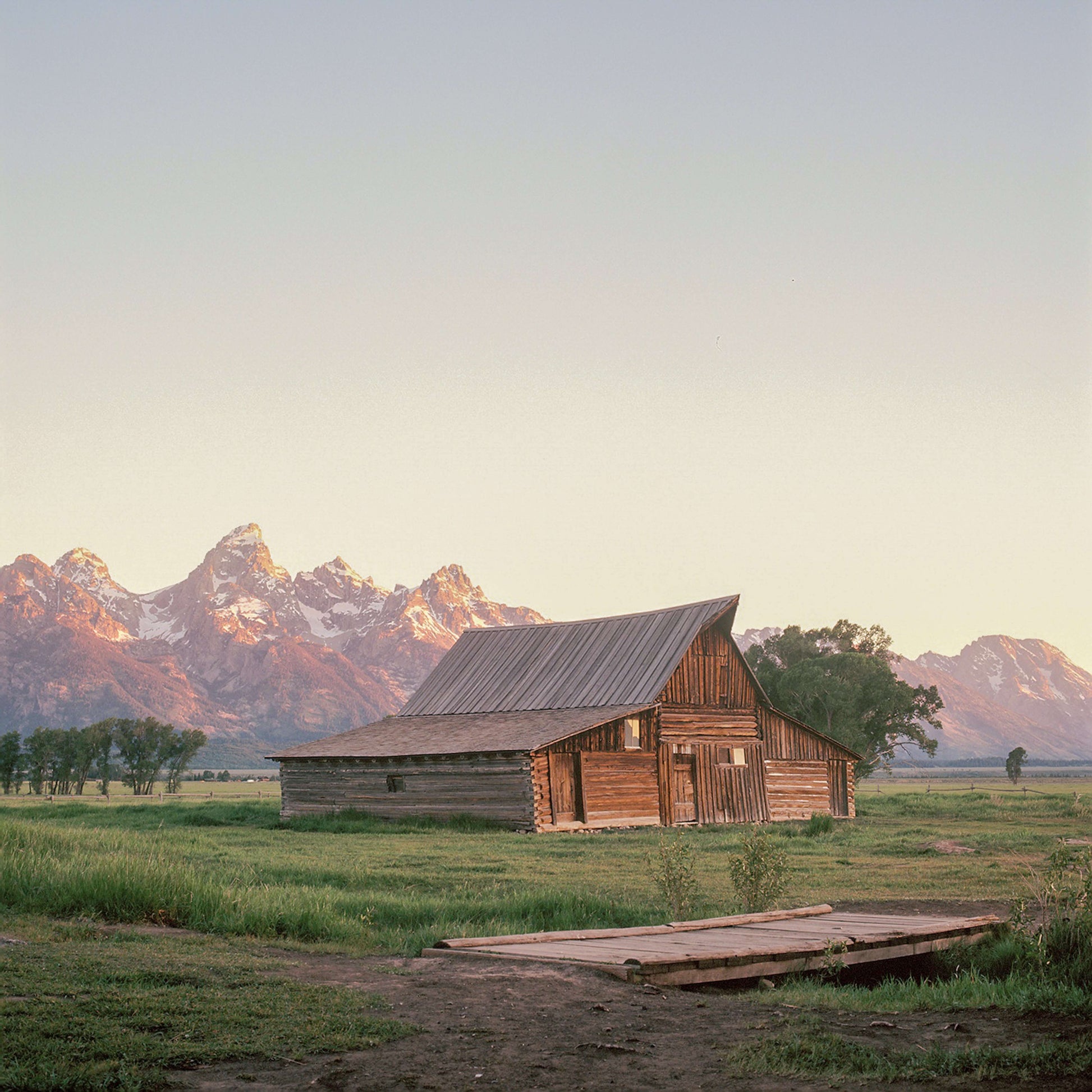 Scenic view of a rustic barn surrounded by lush greenery and mountains at sunrise.