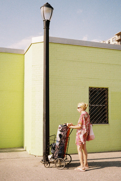 Woman in a floral dress pushing a shopping cart near a bright green wall and a street lamp.