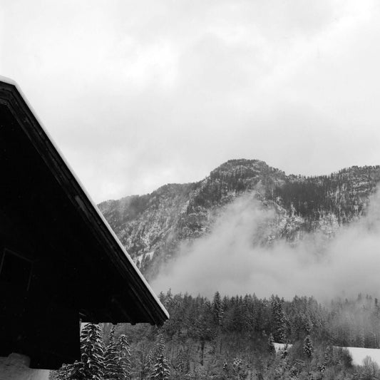Snowy mountain landscape with clouds and trees, featuring the corner of a rustic cabin in black and white.