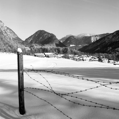 Black and white landscape of a snowy mountain village with barbed wire fence foreground.