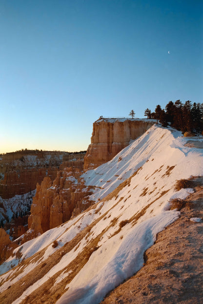 A snowy cliffside in Bryce Canyon with trees and a clear blue sky at sunset.