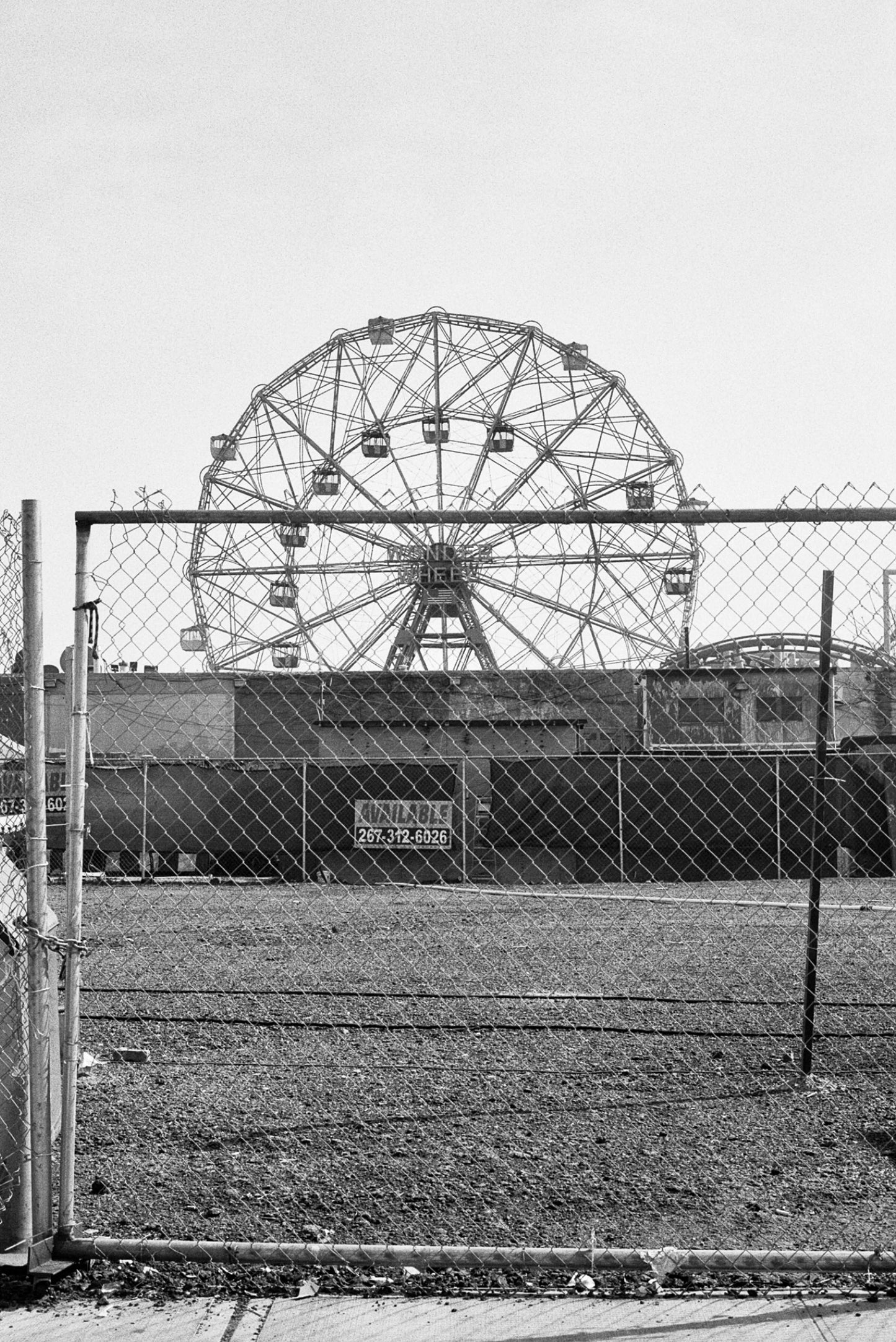Black and white image of a ferris wheel viewed through a fence, capturing a nostalgic amusement park scene.