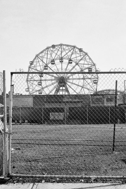 Black and white image of a ferris wheel viewed through a fence, capturing a nostalgic amusement park scene.