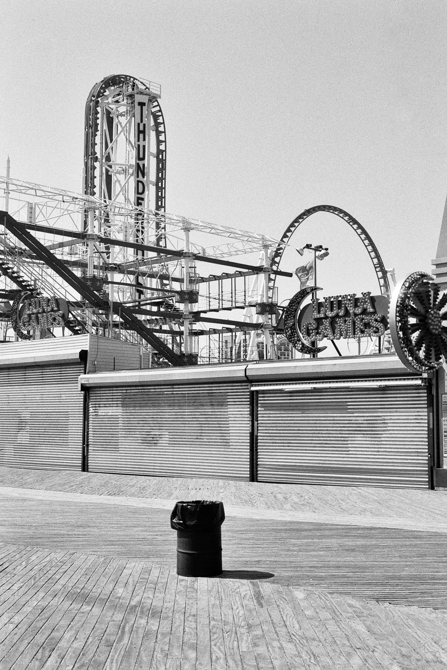 A black and white image of an amusement park with a roller coaster and closed stalls.