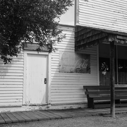 Black and white image of a wooden building with a door and bench, featuring a faded Coca-Cola sign, captured on Ilford Ortho Plus 80.