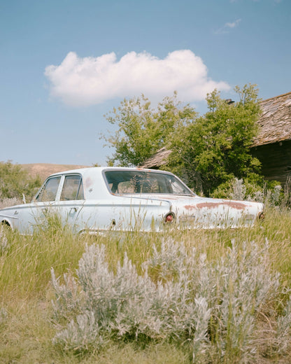 Abandoned blue vintage car in tall grass near old building under a clear sky.