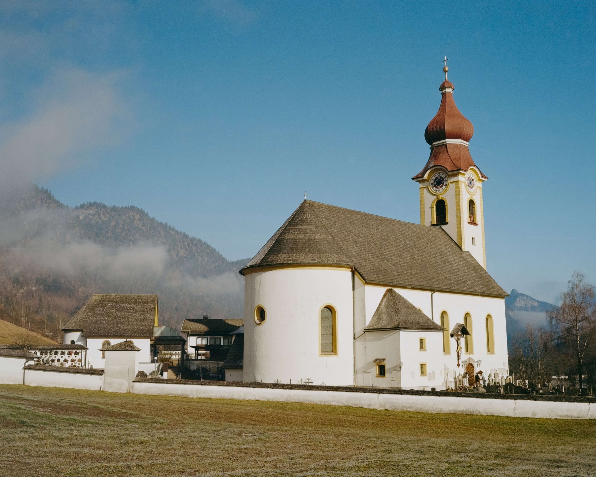 A picturesque church with a red tower and white walls, set against a mountainous backdrop in a serene landscape, shot with Kodak Eastman Gold 200 Color Negative Film