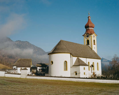 A picturesque church with a red tower and white walls, set against a mountainous backdrop in a serene landscape, shot with Kodak Eastman Gold 200 Color Negative Film