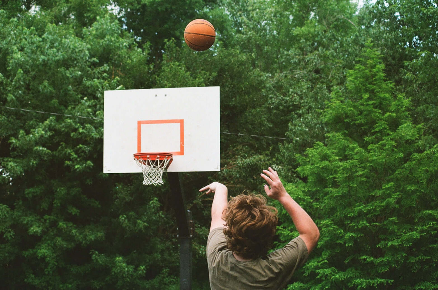 A person shooting a basketball towards a hoop surrounded by lush greenery, shot on Kodak UltraMax 400.