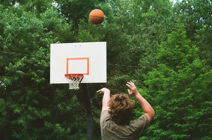 A person shooting a basketball towards a hoop surrounded by lush greenery, shot on Kodak UltraMax 400.