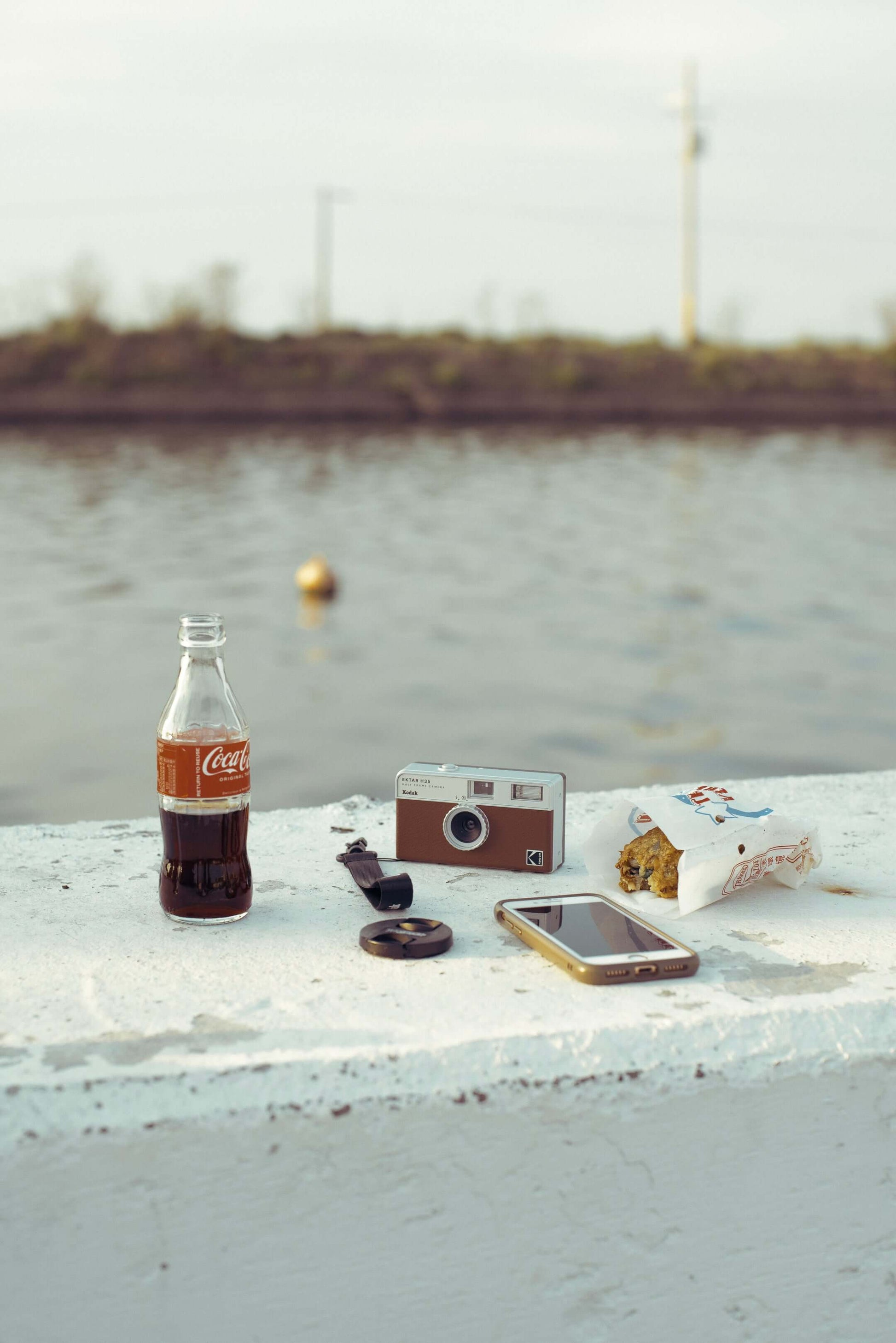 Kodak Ektar H35 Half Frame Film Camera (Brown) on a table with a Coca-Cola and a smartphone by the water.