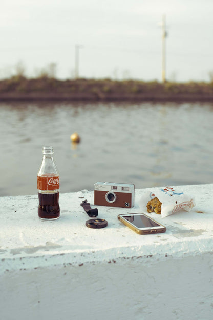Kodak Ektar H35 Half Frame Film Camera (Brown) on a table with a Coca-Cola and a smartphone by the water.