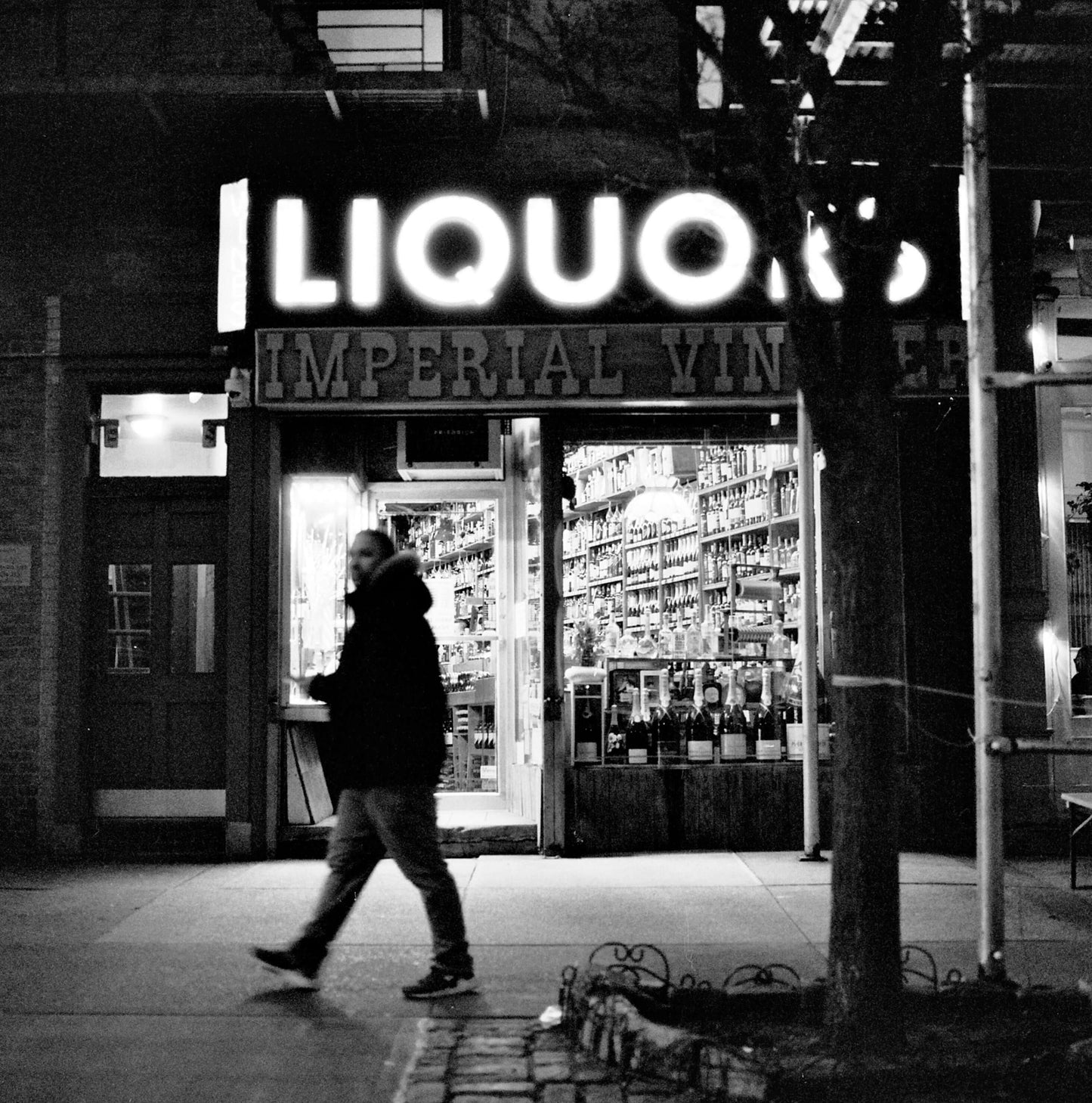 A man walking past an illuminated liquor store at night, showcasing the vibrant signs and bottle displays inside, captured on Ilford Delta 3200.