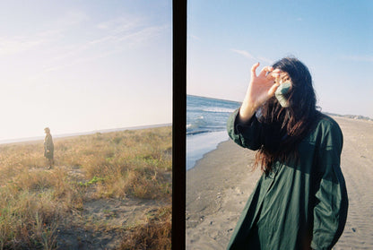 Person standing on a sandy beach, split by a vertical line with sea and sky on the left and a woman in a green coat sipping from a cup on the right, shot using Ektar H35 Half Frame camera.