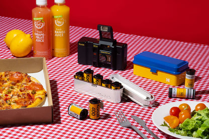Kodak 135 Film Case (Silver) with 35mm film rolls, pizza, and drinks on a picnic table.