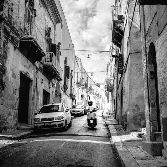narrow street scene with parked cars and a scooter in black and white, showcasing urban architecture and cloudy sky shot with Kentmere Pan 100 Black and White Negative Film.