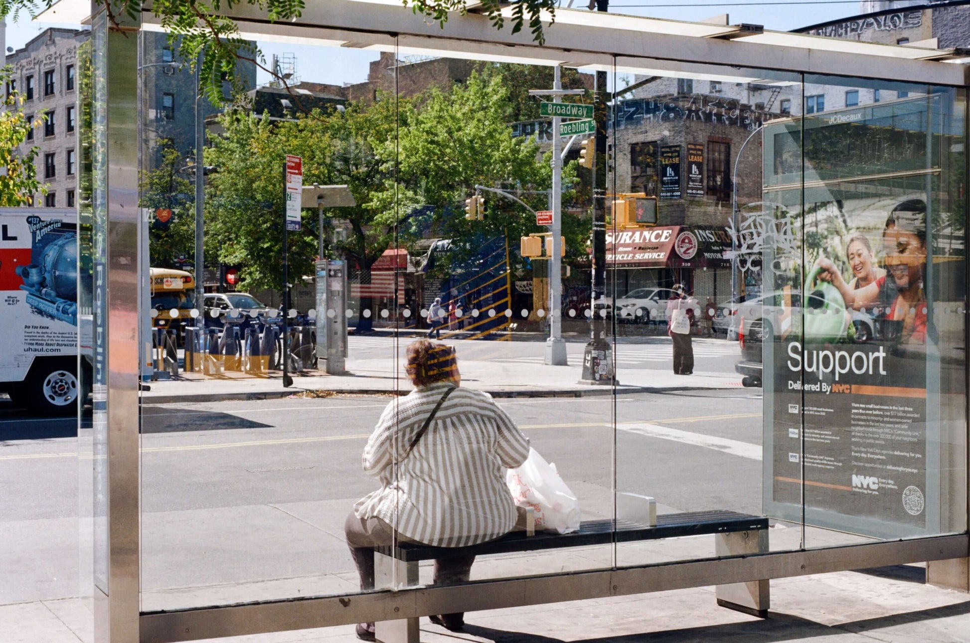 Person sitting at a bus stop with a transparent shelter on a sunny day in an urban setting, shot on Kodak Kodacolor 100.