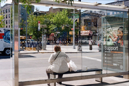Person sitting at a bus stop with a transparent shelter on a sunny day in an urban setting, shot on Kodak Kodacolor 100.