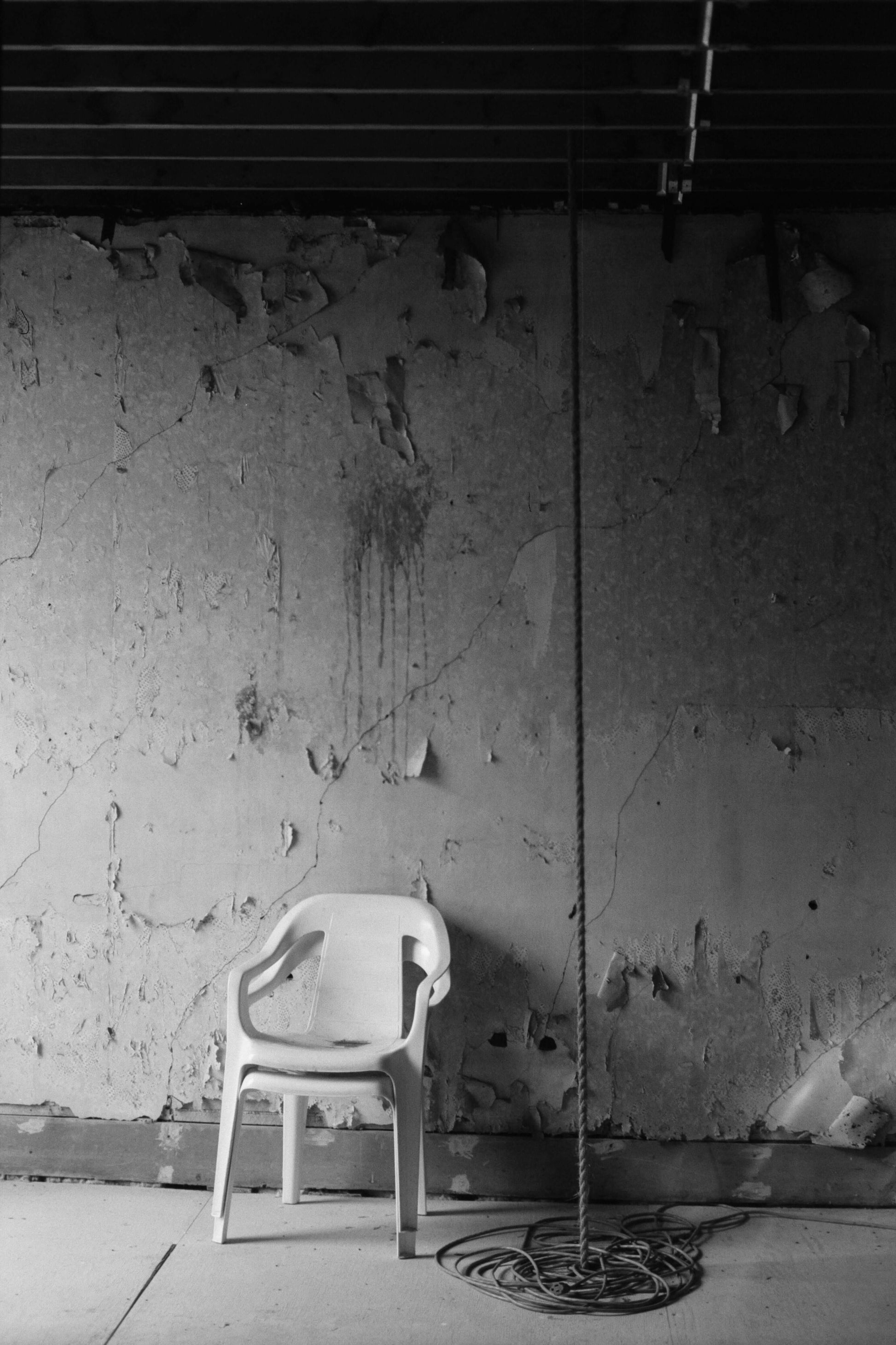 A solitary plastic chair in front of an aged, textured wall with a rope hanging nearby, captured in black and white shot with Mr Negative Cash Only 100