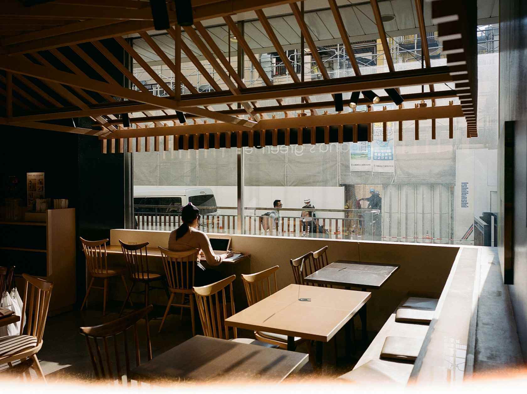 Interior of a cafe with a woman sitting at a table, bright natural light streaming through large windows.