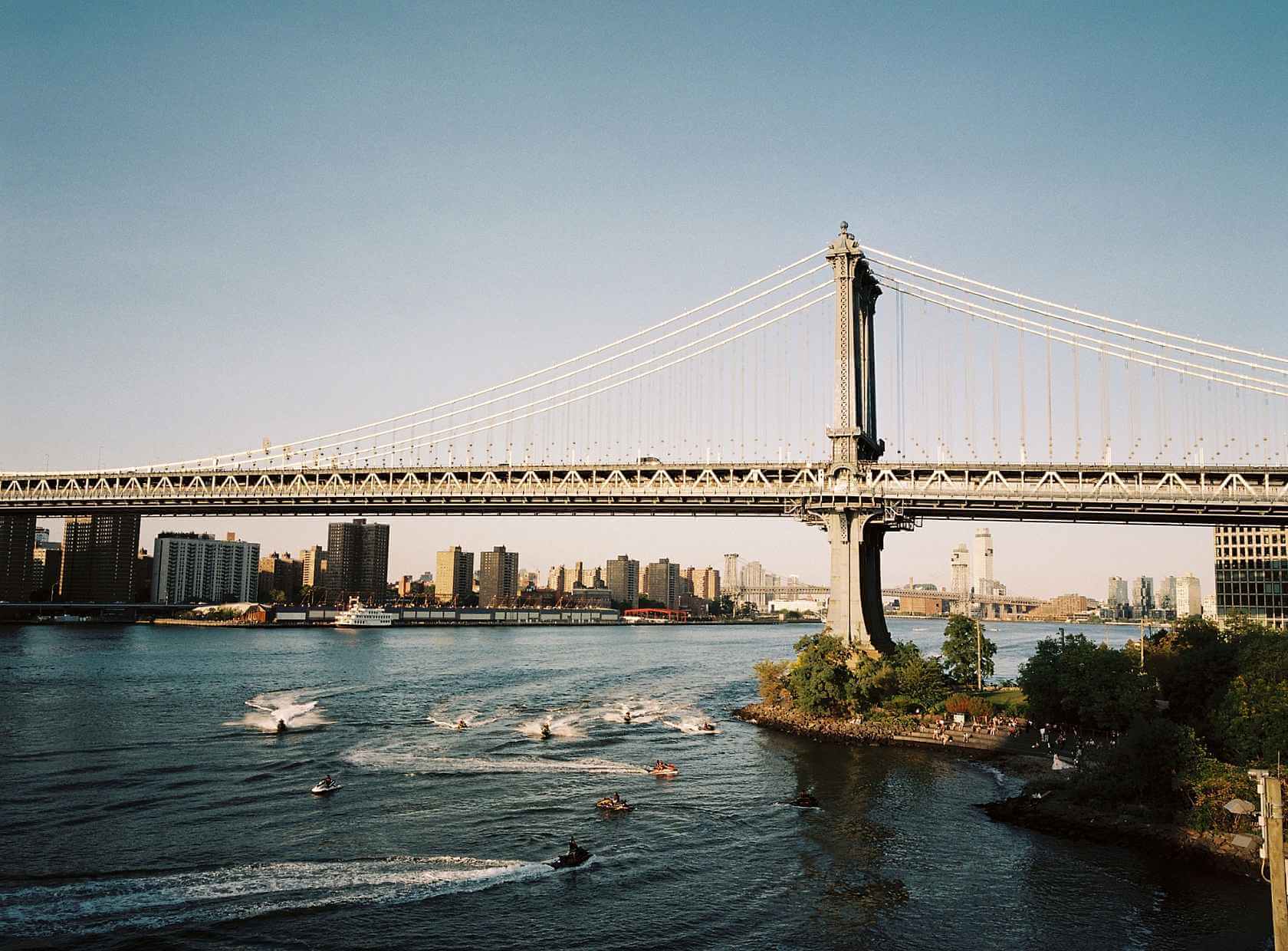 Manhattan Bridge spanning the East River with boats and city skyline in view, under a clear blue sky.