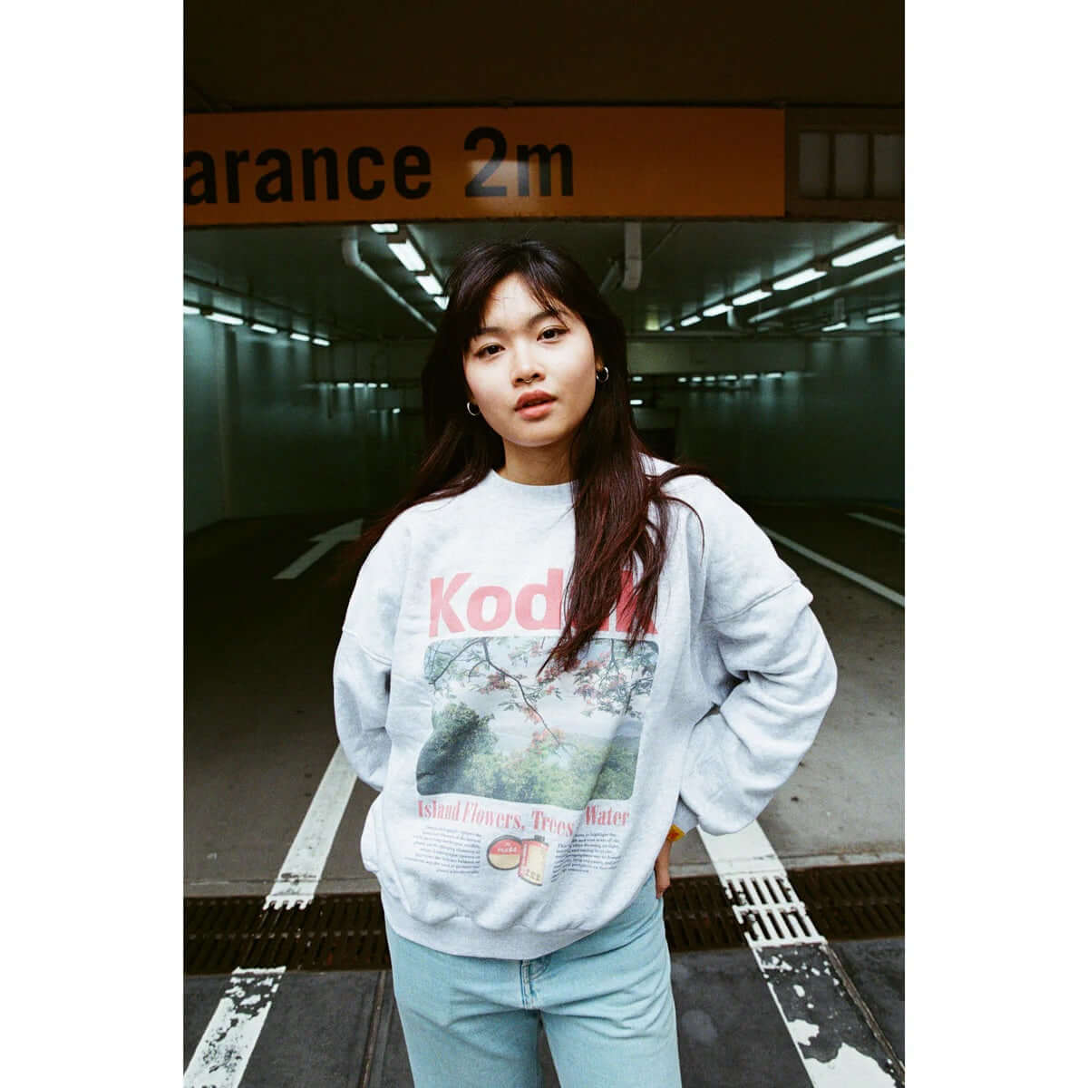 A woman wearing a Kodak sweatshirt stands in a parking garage with a 2m clearance sign.