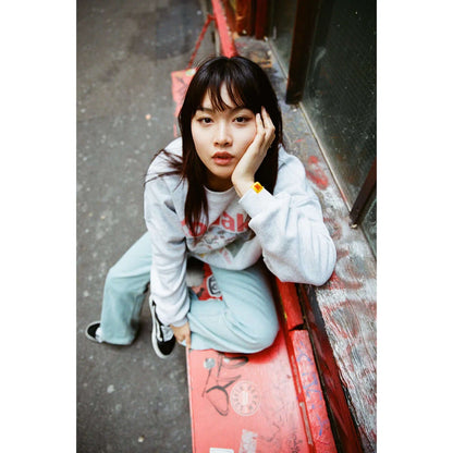 Model wearing a gray sweatshirt and light blue pants, sitting on a red bench in an urban setting.